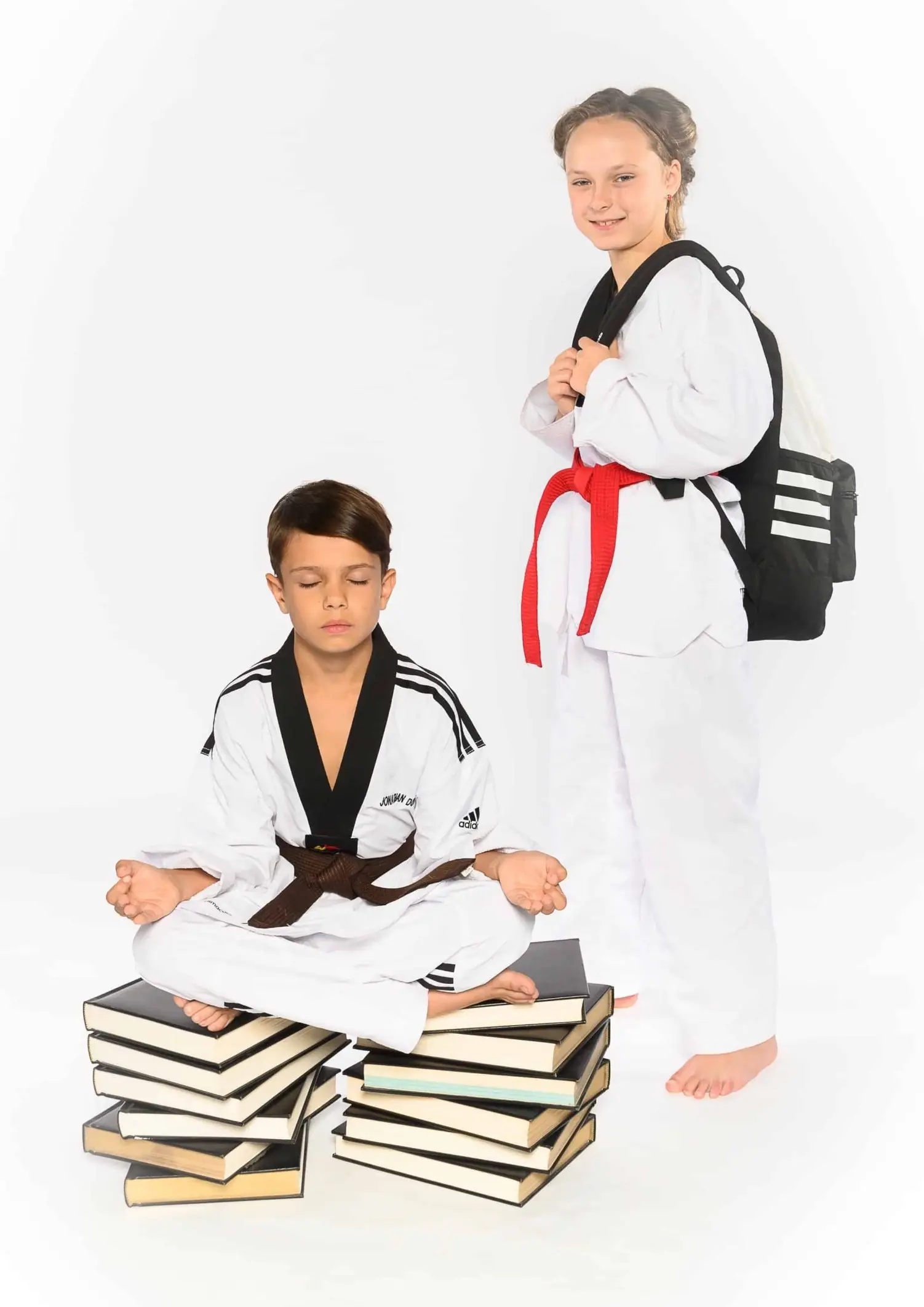 Two Dragon Taekwondo students in uniform, one sitting on a stack of books and one wearing a school backpack