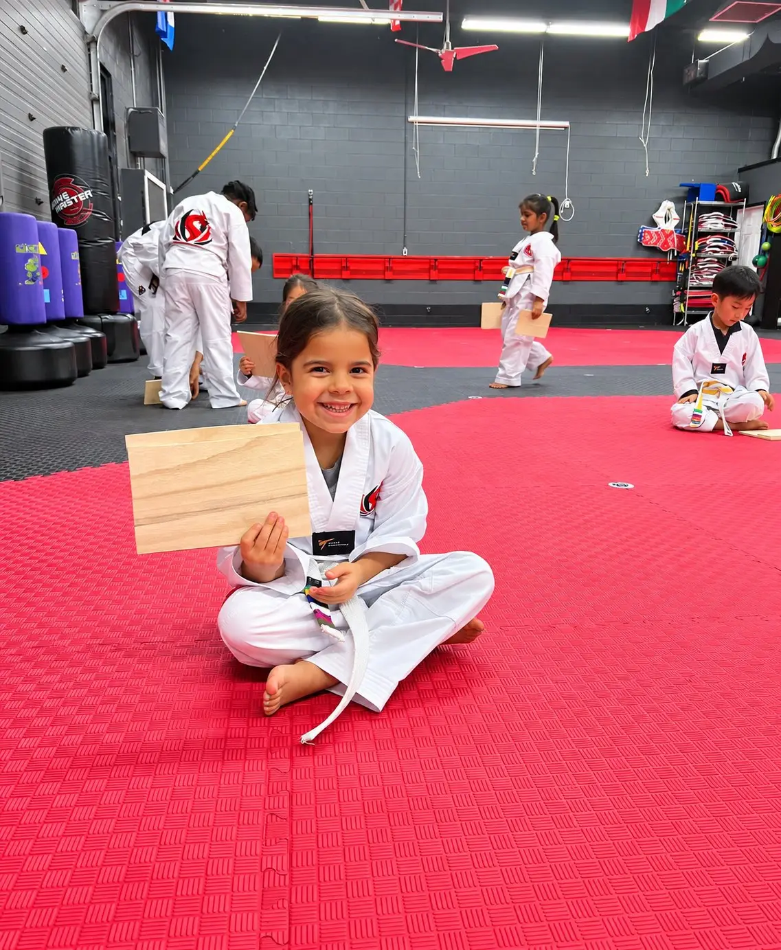 A smiling Little Dragons student sitting on the mat holding the board she broke