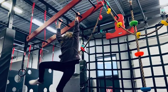 A child on the monkey bars of the indoor ninja course, next to a cargo net and climbing holds